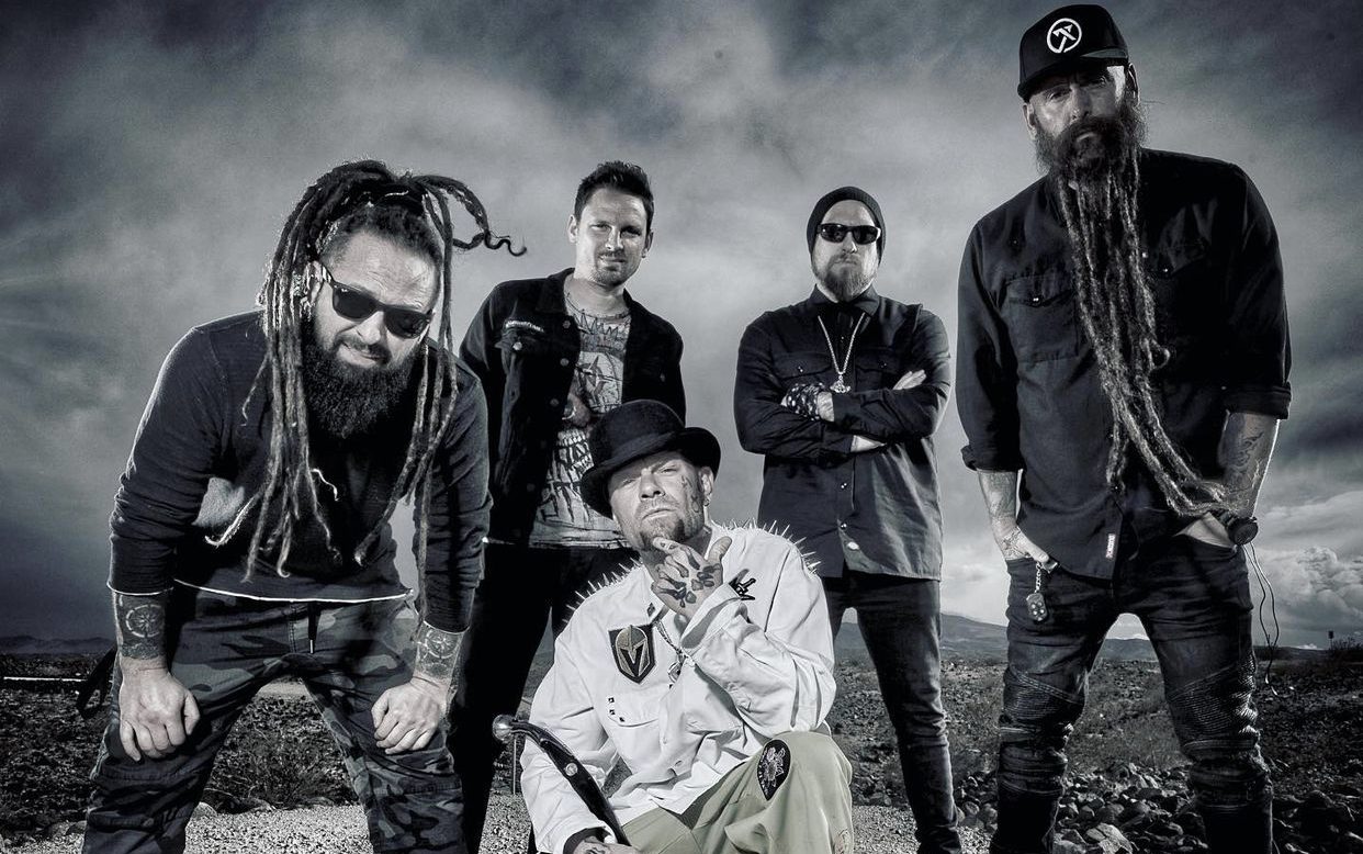 Members of Five Finger Death Punch stand behind member who is squatting in a white shirt. They are outside in front of a dim sky.