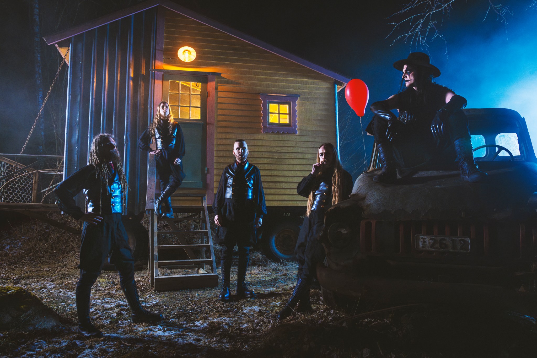 Members of Avatar in front of a cabin at night. The vocalist is sitting on an old truck and a red balloon is next to him.