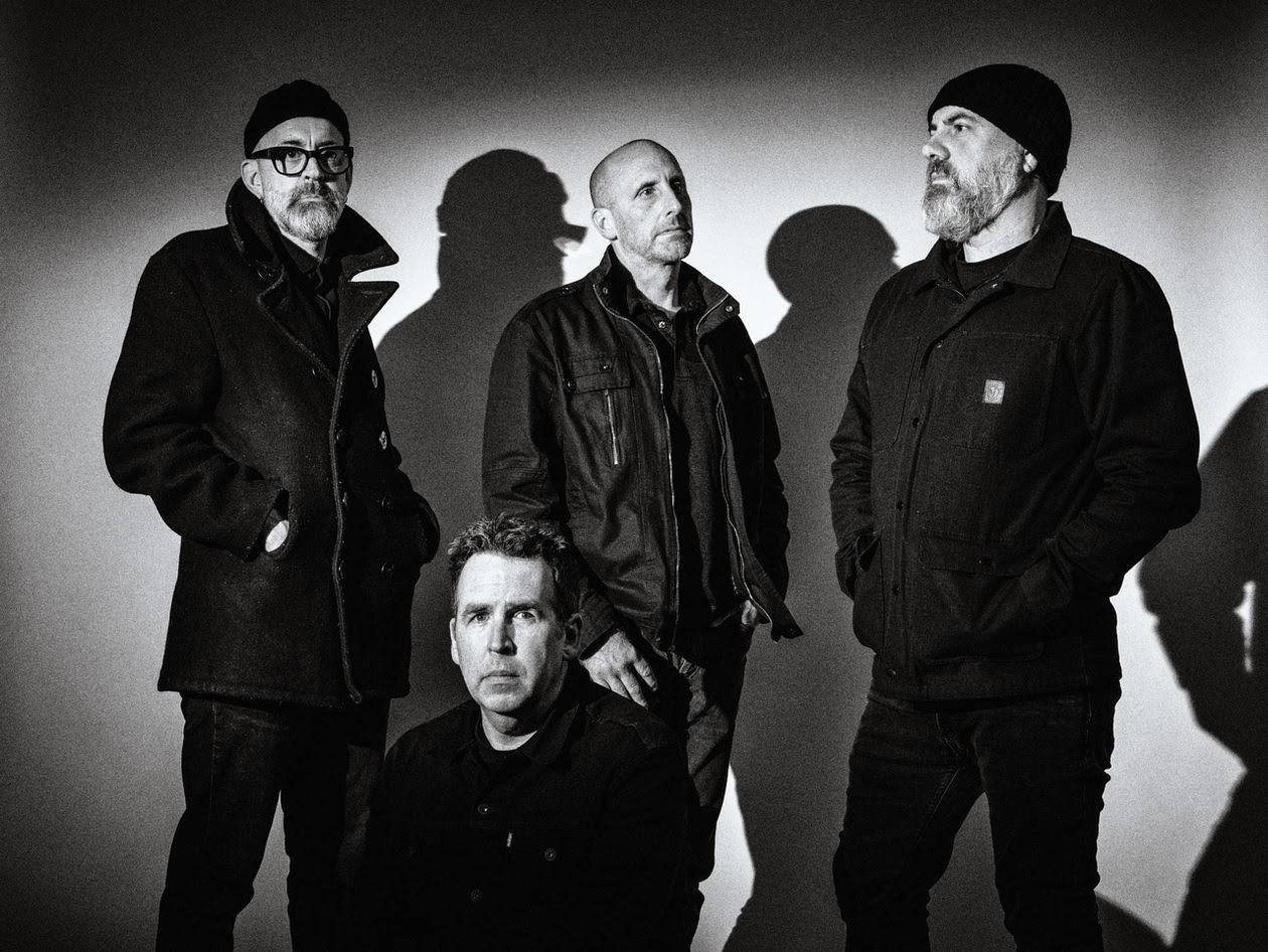 Band all dressed in black standing together, one squatted toward the front. Black and white photo with dark edges.