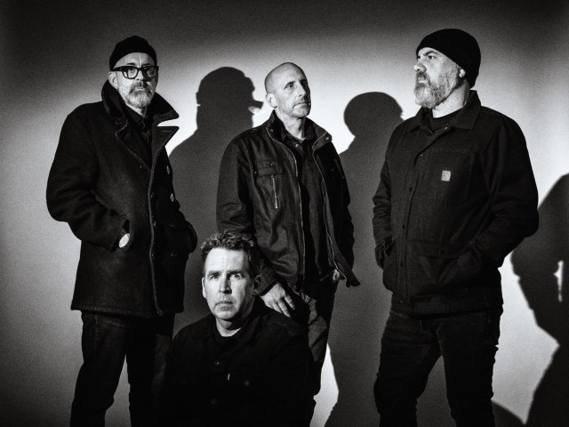Band all dressed in black standing together, one squatted toward the front. Black and white photo with dark edges.