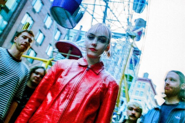 Members of Just Mustard stand in front of a ferris wheel, the lead singer wears a red jacket.
