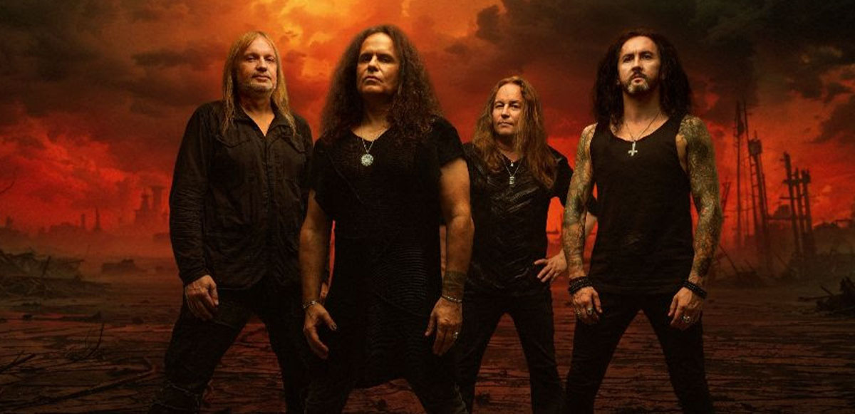 Kreator members wearing all black standing in front of a sea with a shipwreck in the background and red sky.