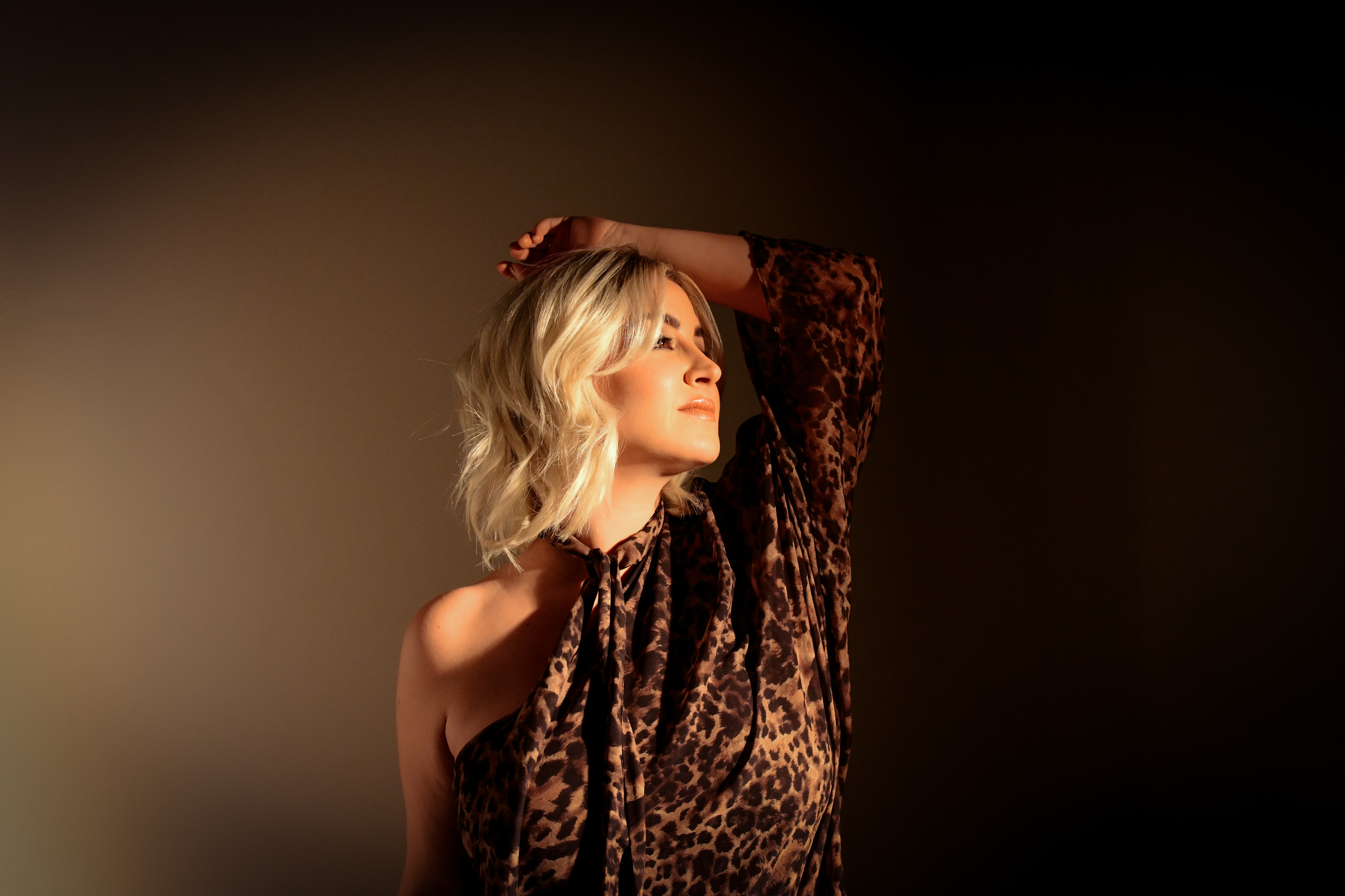 Maggie Rose stands with her head over her head facing right in front of a brown background.