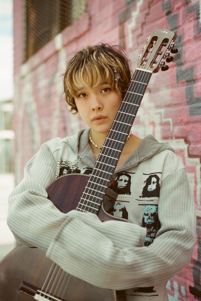 Mei Semones standing in front of a pink wall outside while holding an acoustic guitar.