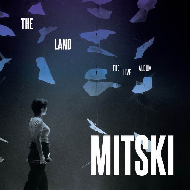 Black and white photo of Mitski standing, facing away from the camera, while blue and purple specs fall from the top left.