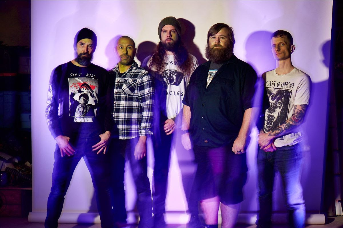 Band members stand together in front of a blank wall lit by a purple light
