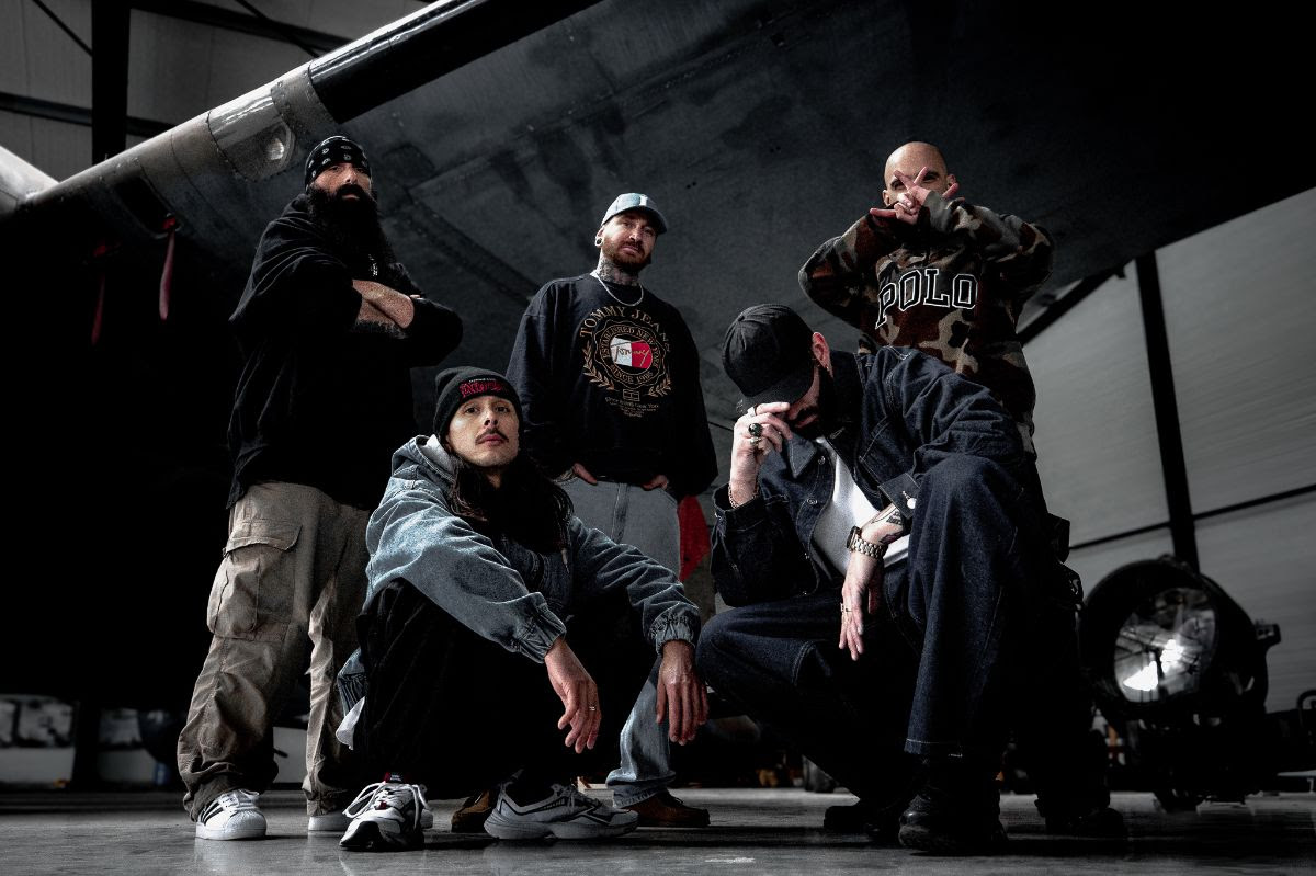 Band members stand together in a hangar, two squatted on the floor in front of the others.
