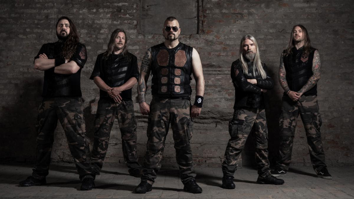 Members of Sabaton standing in front of a stone wall, the frontman wearing an armor vest