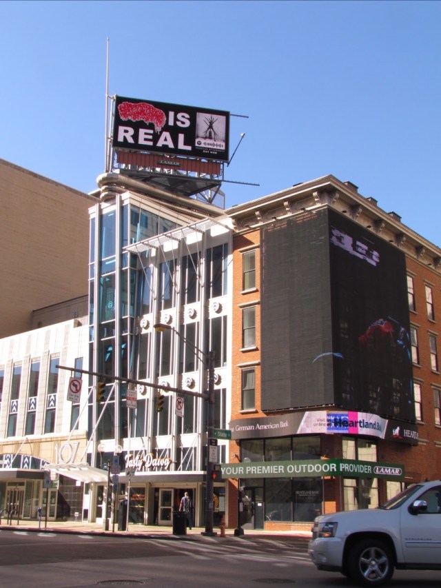Billboard with "[Band Logo] IS REAL" next to album art over a tall building.