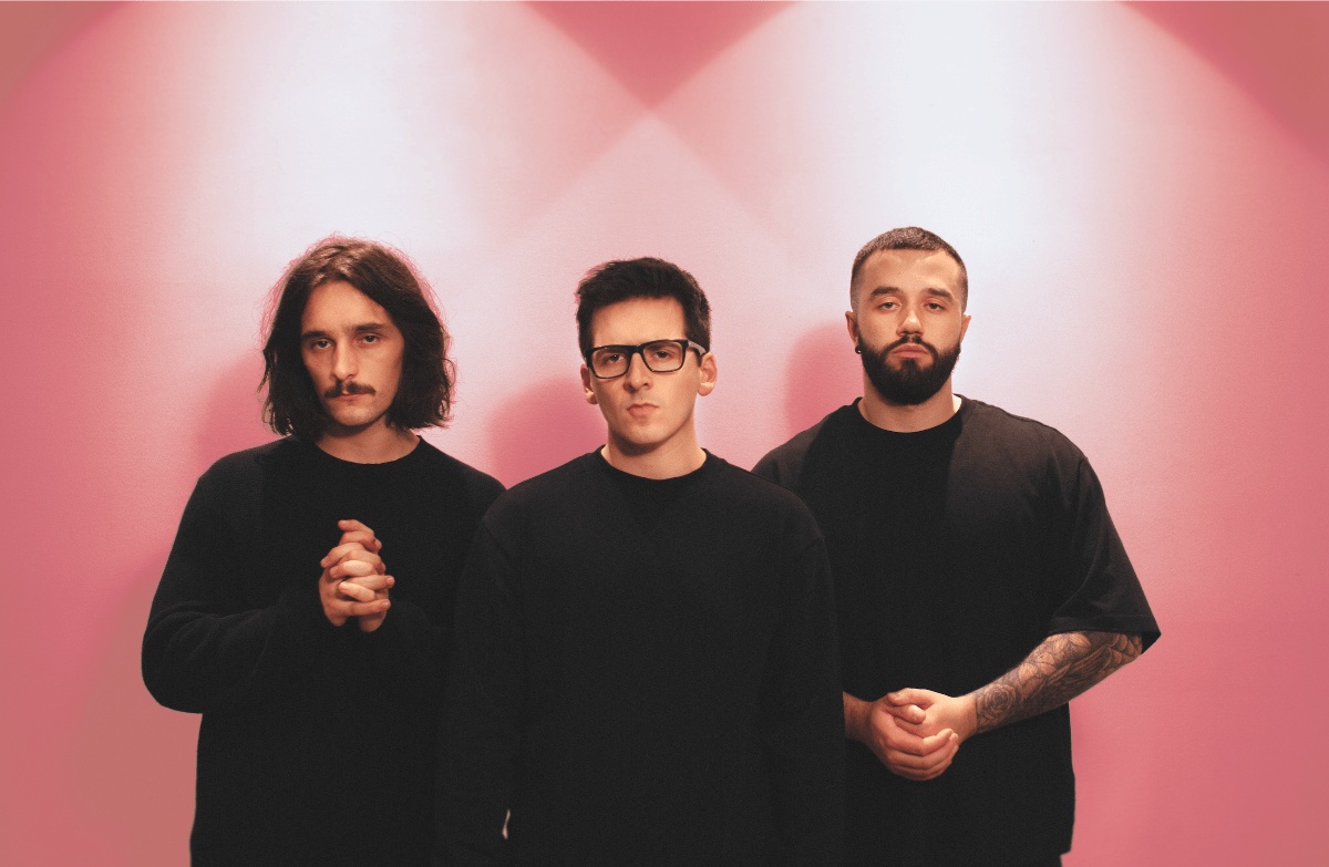 Band members stand in front of a pink wall while wearing all black.