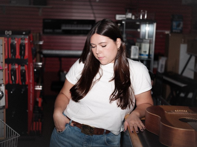 Gigi Perez looking down and to the side next to a guitar that is sitting on a table.