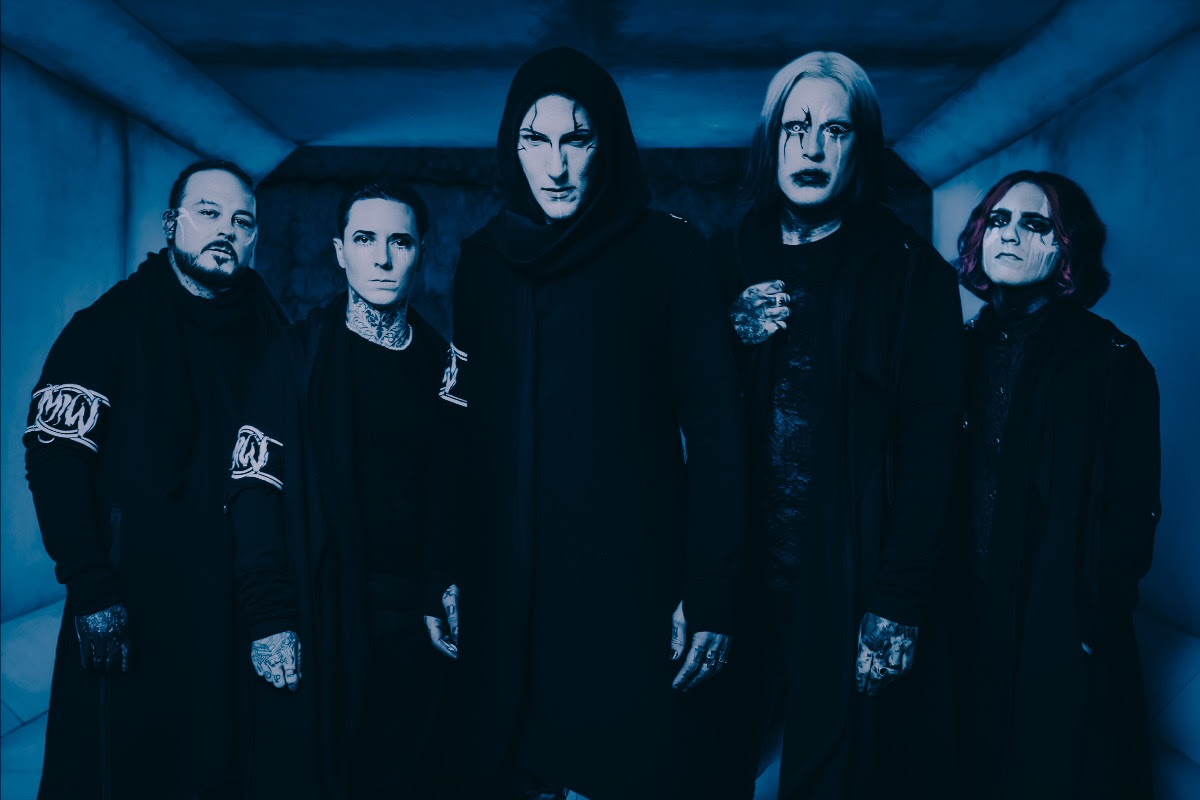 Motionless In White members standing in a dark hall way all dressed in black. A blue filter is put over the photo.
