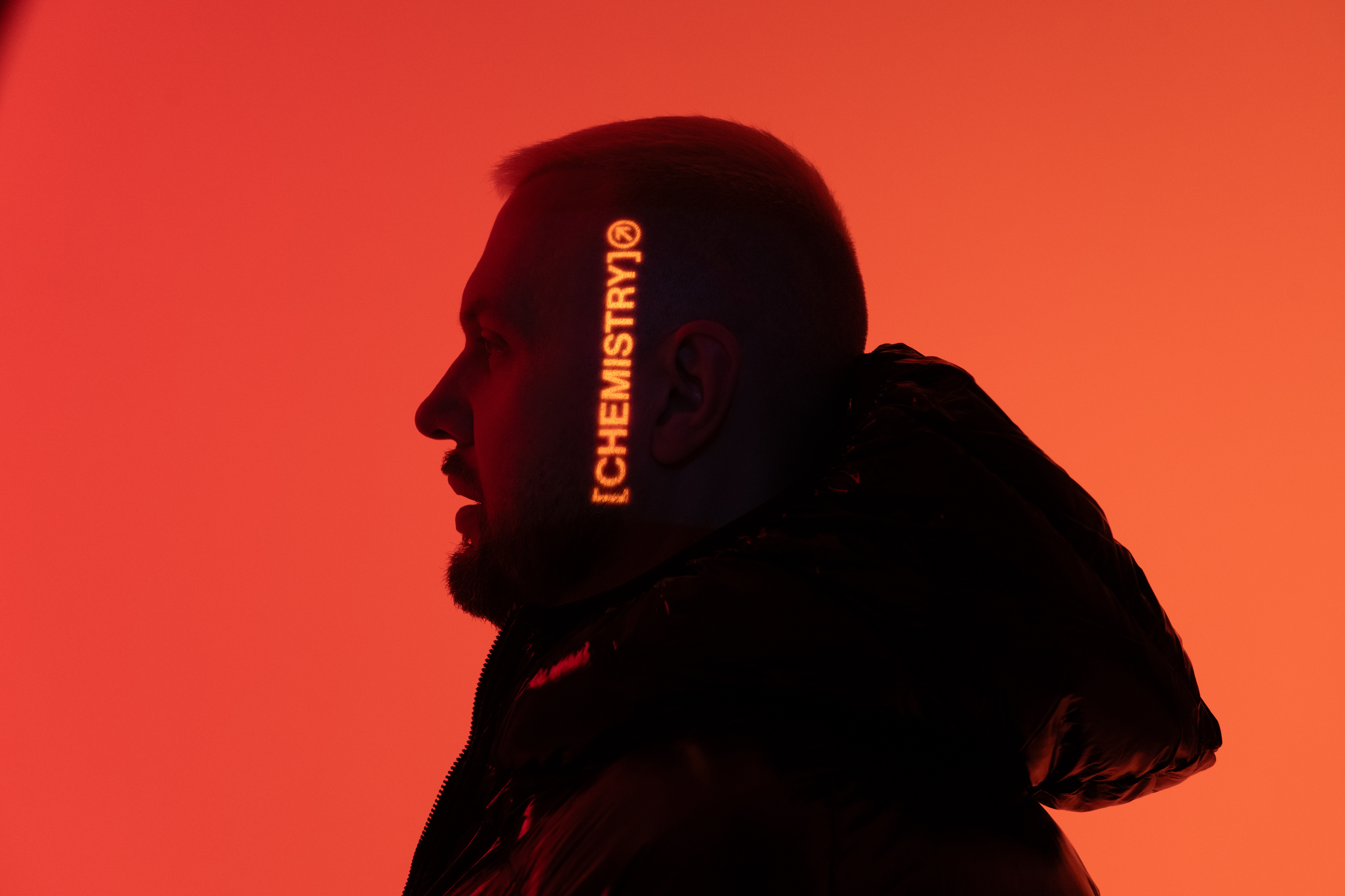 Chris Lake standing in front of an orange background with the word "Chemistry" projected onto his head.