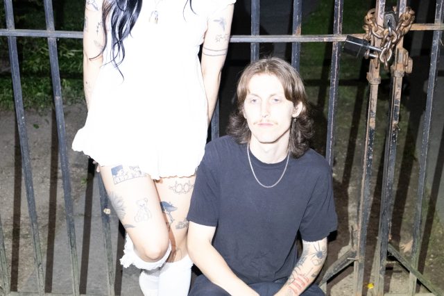 Memers of smush in front of a metal fence at night with the camera flash on. The singer is wearing a white dress and seen from the shoulders down while the guitarist is sitting in a black shirt.
